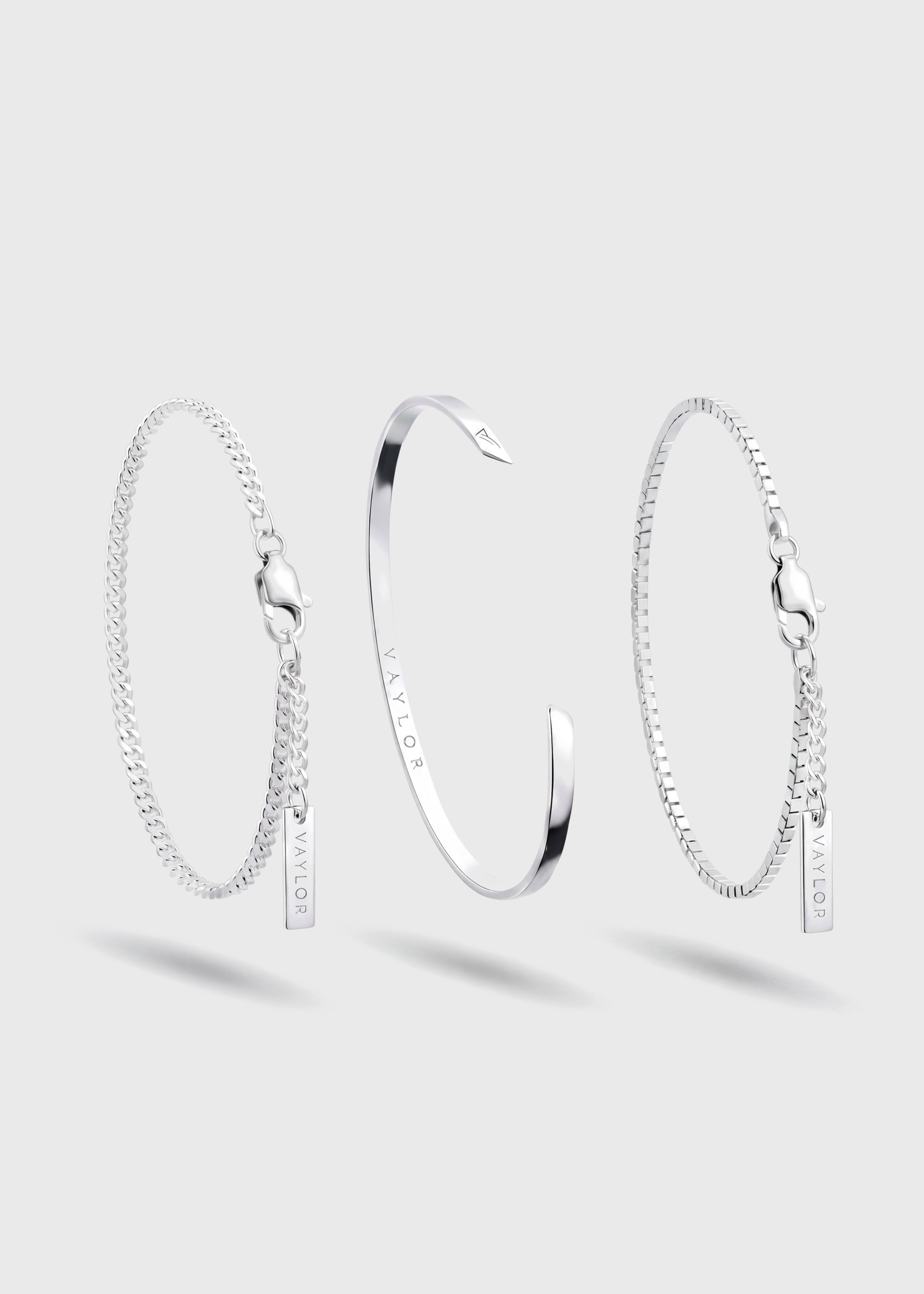 Three silver chain bangles displayed on a light gray background: a textured chain bracelet with a small hanging tag on the left, a polished open cuff bangle in the center, and a textured chain bracelet with a small hanging tag on the right.