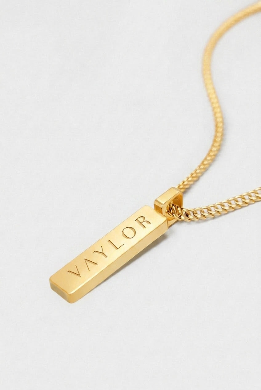 Gold necklace with a bar pendant engraved with 'VAYLOR' on a light gray background