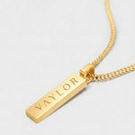 Gold necklace with a bar pendant engraved with 'VAYLOR' on a light gray background