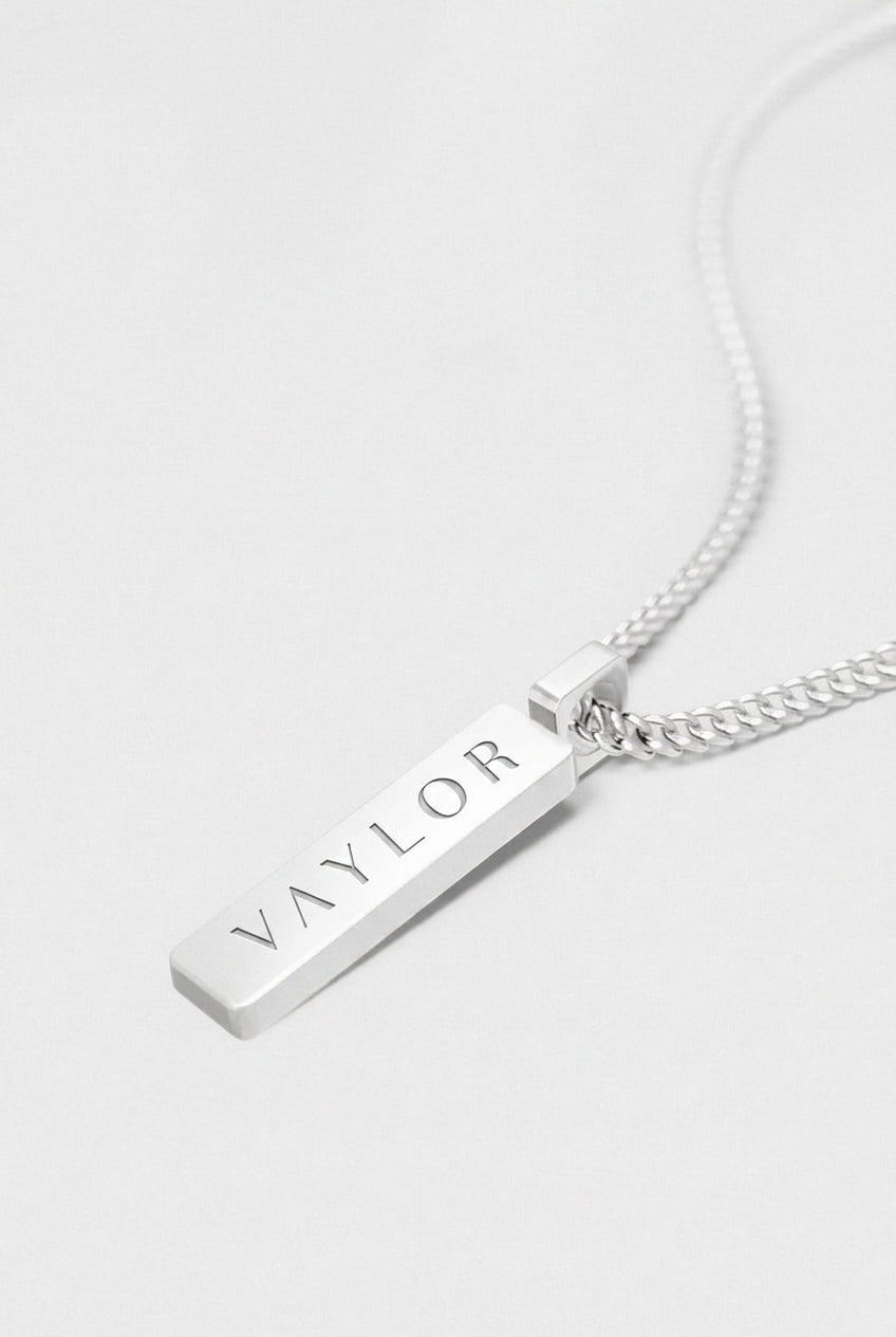 Silver necklace with a bar pendant engraved with 'VAYLOR' on a light gray background