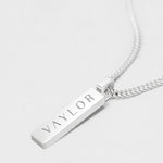 Silver necklace with a bar pendant engraved with 'VAYLOR' on a light gray background
