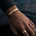 Hand wearing gold bracelets and a ring with water in the background