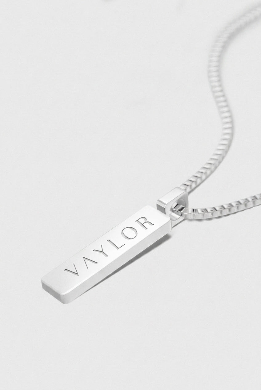 Silver necklace with a bar pendant engraved with 'VAYLOR' on a white background