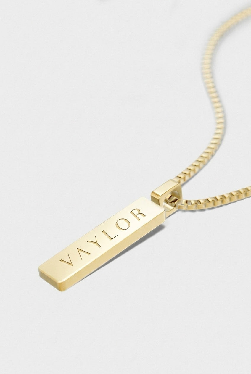 Gold necklace with a bar pendant engraved with 'VAYLOR' on a light gray background