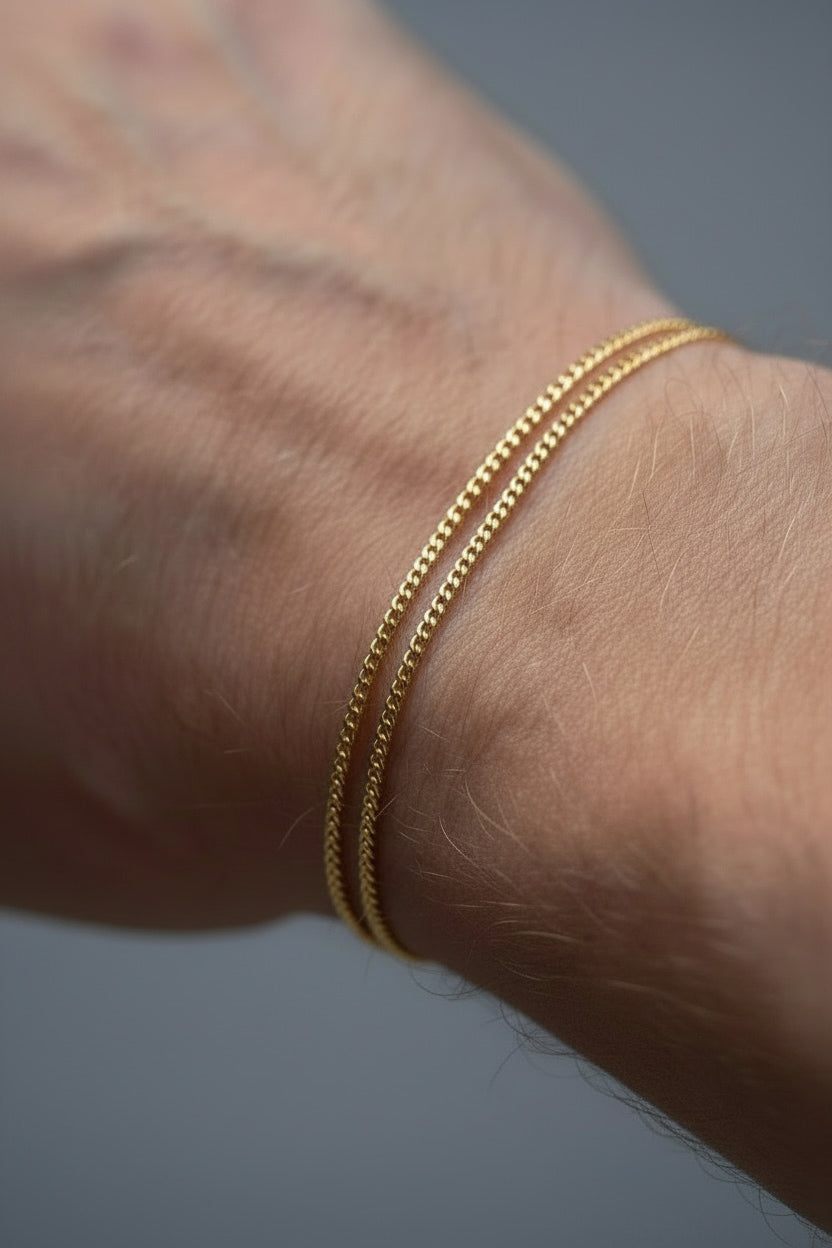 Two thin gold chain bracelets worn together on a person's wrist, shown in a close-up shot against a neutral gray background.