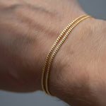 Two thin gold chain bracelets worn together on a person's wrist, shown in a close-up shot against a neutral gray background.