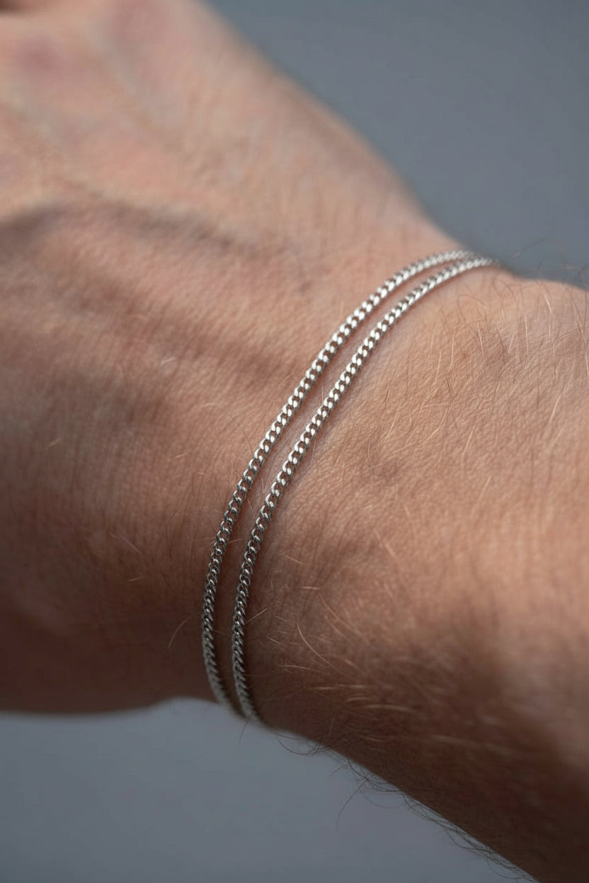 Two thin silver chain bracelets worn on a person's wrist against a soft gray background.