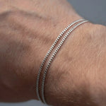 Two thin silver chain bracelets worn on a person's wrist against a soft gray background.
