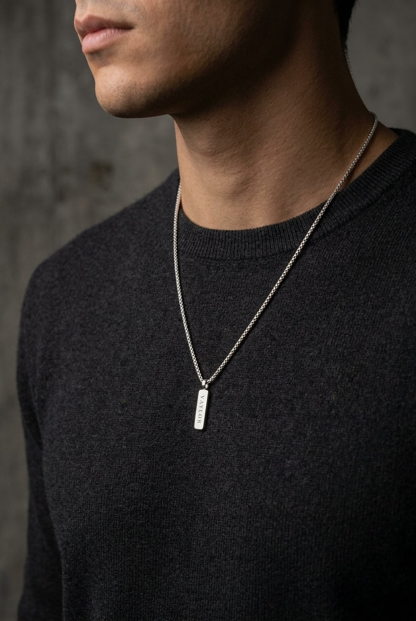 Man wearing a black sweater with a silver necklace featuring a rectangular pendant against a dark background