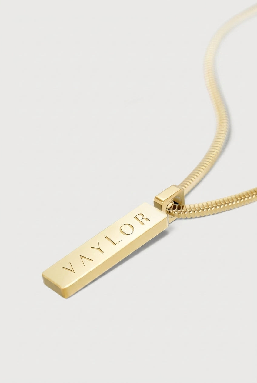 Gold necklace with a bar pendant engraved with 'VAYLOR' on a light gray background