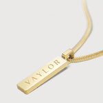 Gold necklace with a bar pendant engraved with 'VAYLOR' on a light gray background