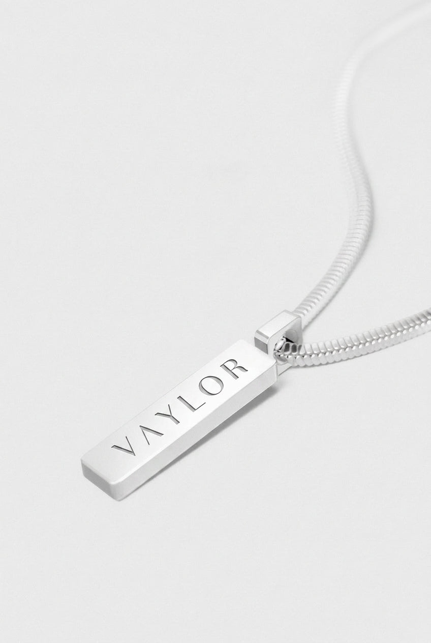 Silver necklace with a bar pendant engraved with 'VAYLOR' on a white background