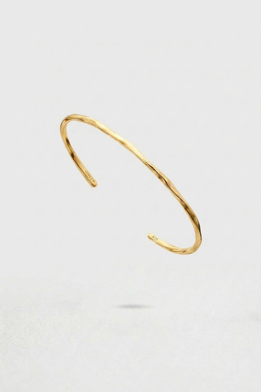 A thin, open cuff bangle bracelet with a hammered gold finish, shown floating against a light gray background.