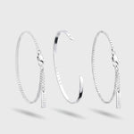 Three silver chain bangles displayed on a light gray background: a textured chain bracelet with a small hanging tag on the left, a polished open cuff bangle in the center, and a textured chain bracelet with a small hanging tag on the right.