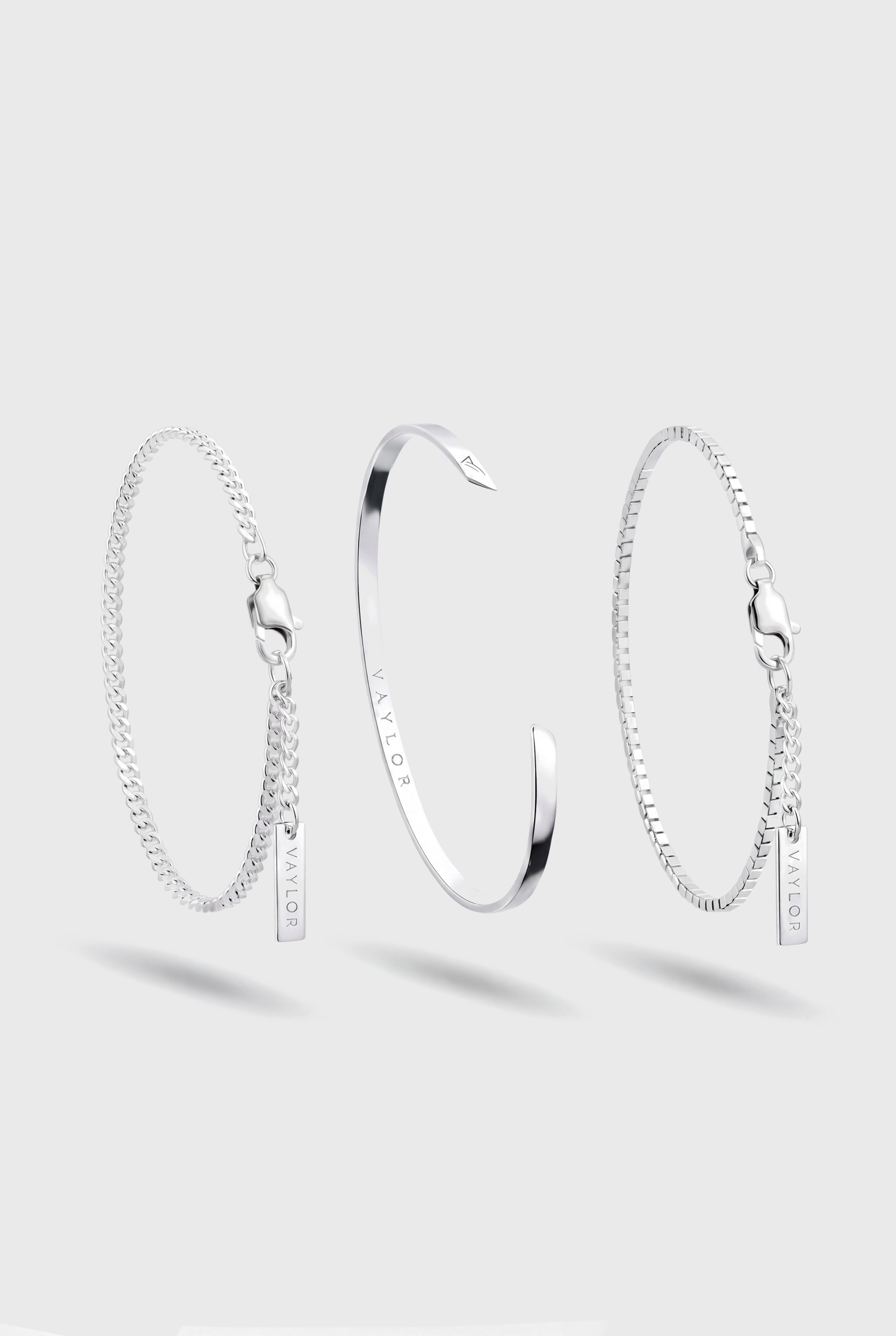 Drie zilveren schakelarmbanden op een lichtgrijze achtergrond: een getextureerde schakelarmband met een klein hangertje links, een gepolijste open manchetarmband in het midden, en een getextureerde schakelarmband met een klein hangertje rechts.