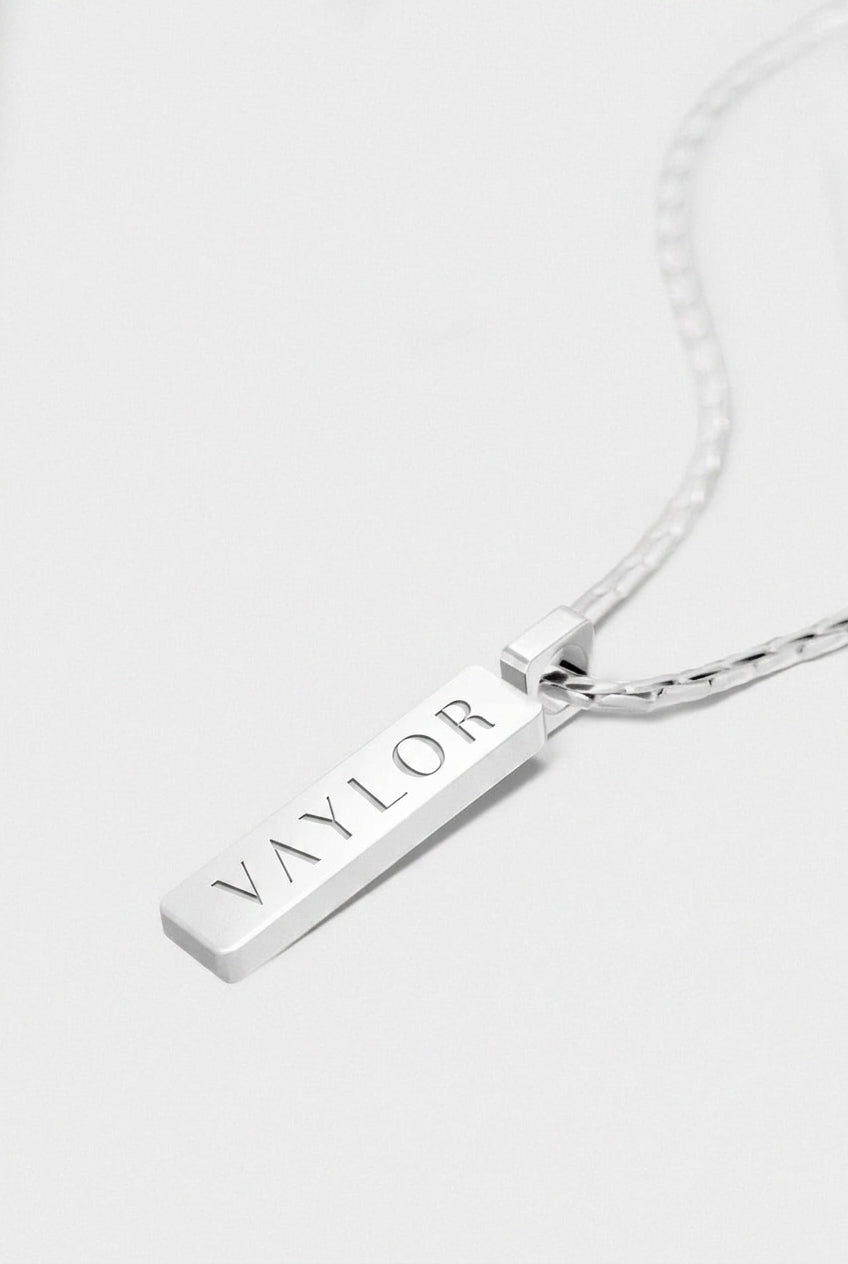 Silver necklace with a bar pendant engraved with 'VAYLOR' on a white background
