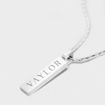 Silver necklace with a bar pendant engraved with 'VAYLOR' on a white background