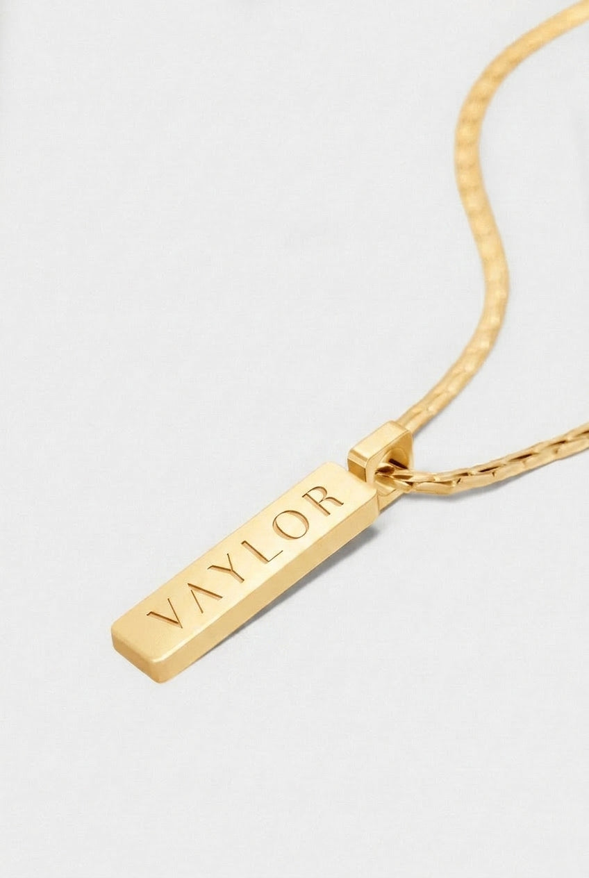 Gold necklace with a bar pendant engraved with 'VAYLOR' on a light gray background