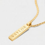 Gold necklace with a bar pendant engraved with 'VAYLOR' on a light gray background