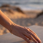A close-up of a person's hand resting on a stone surface, wearing a delicate gold chain bracelet, with a blurred beach and ocean background.