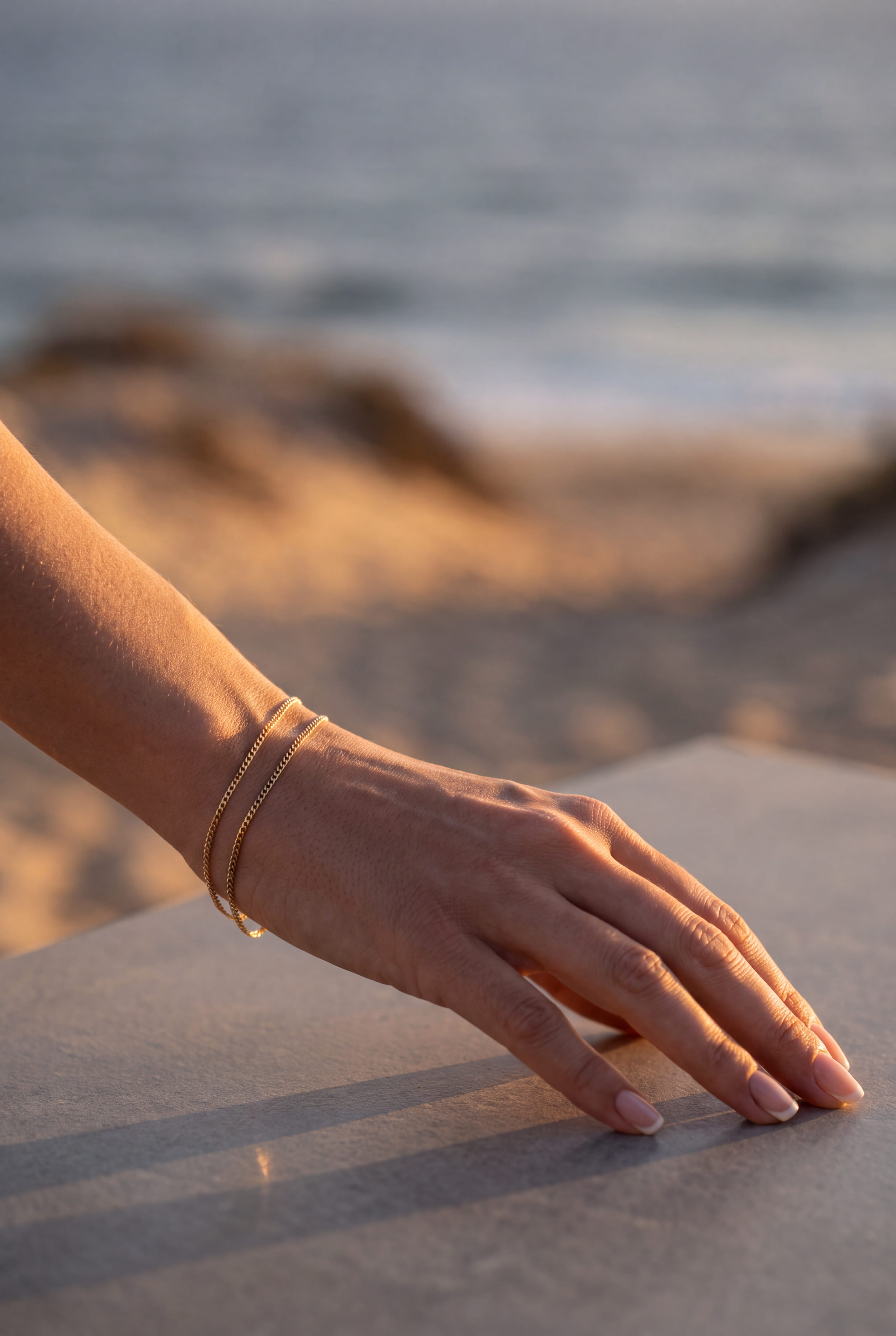 A close-up of a person's hand resting on a stone surface, wearing a delicate gold chain bracelet, with a blurred beach and ocean background.
