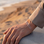 A close-up of a person's hand resting on a concrete ledge at a beach, wearing two delicate silver chain bracelets.