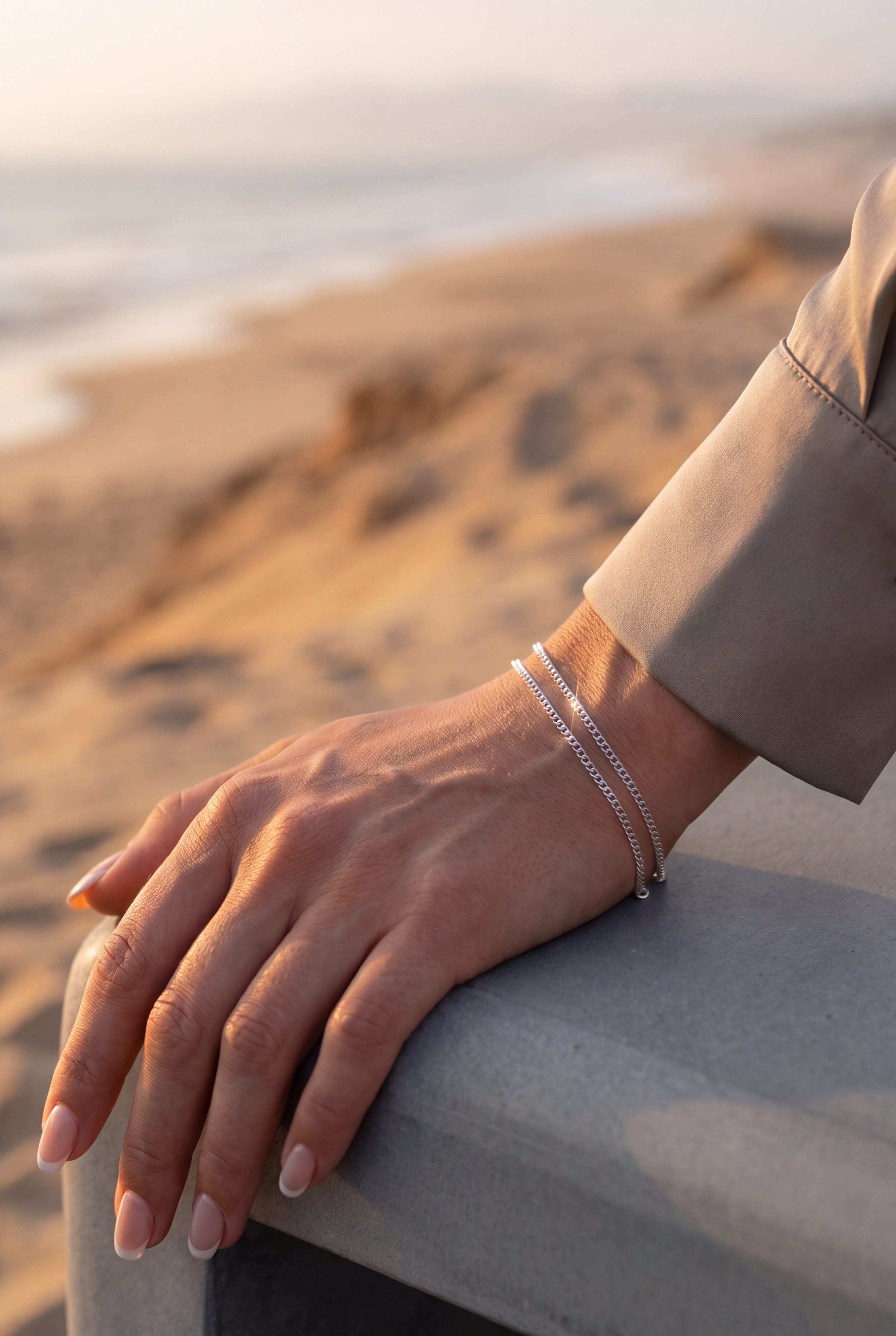 A close-up of a person's hand resting on a concrete ledge at a beach, wearing two delicate silver chain bracelets.