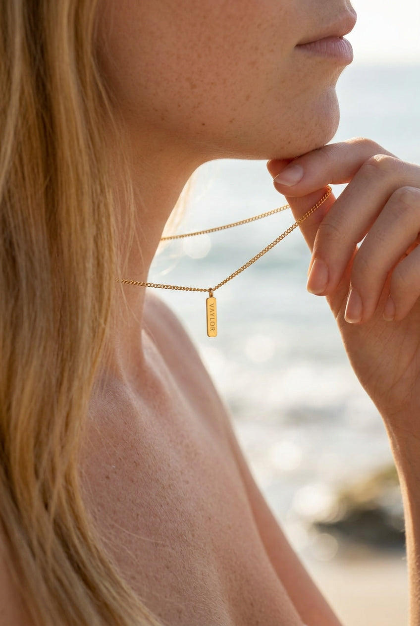 Close-up of a person wearing a gold necklace with a pendant, blurred beach background