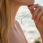Close-up of a person wearing a gold necklace with a pendant, blurred beach background