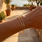 Hand wearing a gold bracelet with a scenic background of a stone building and ocean.