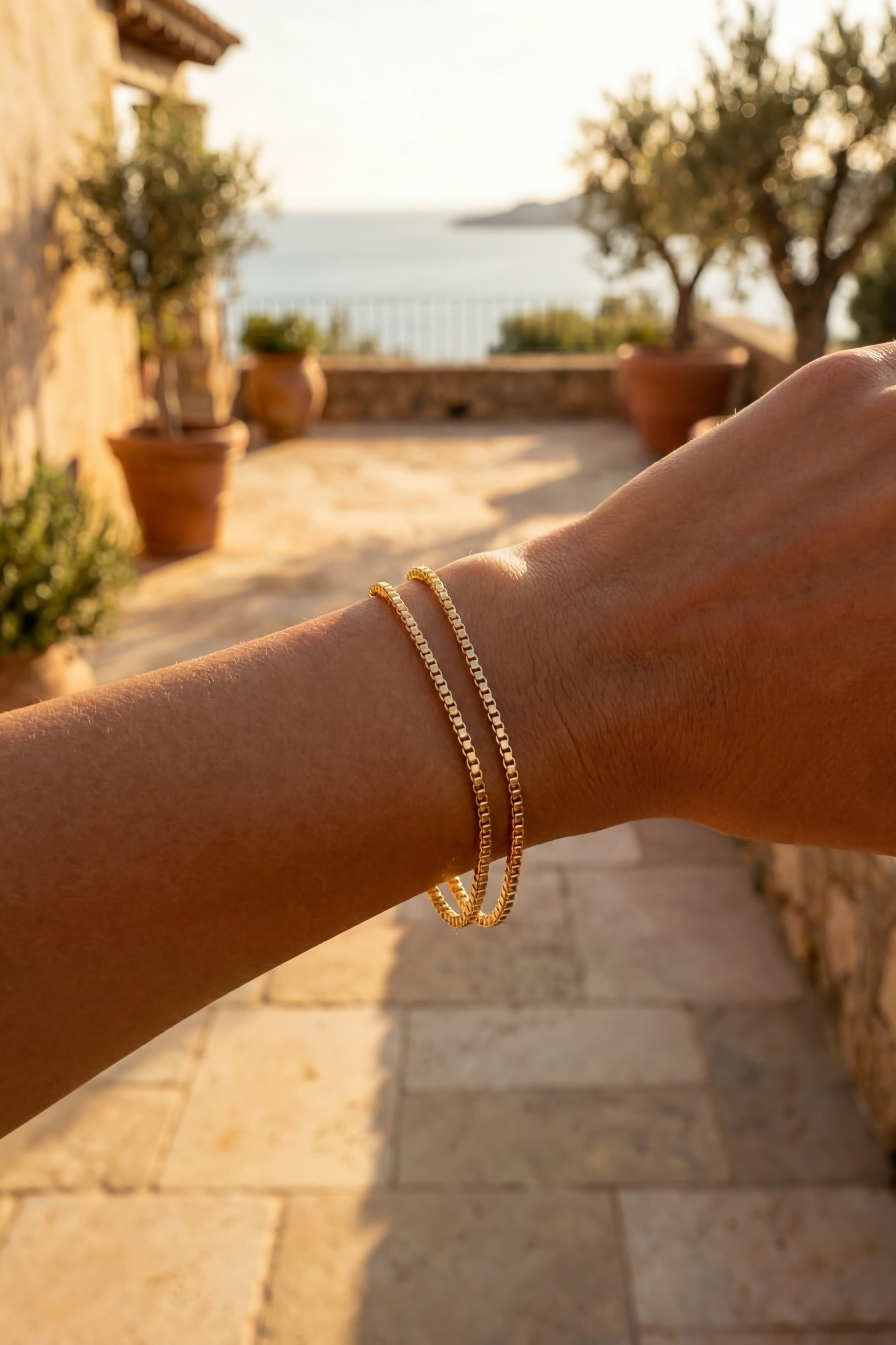 Hand wearing a gold bracelet with a scenic background of a stone building and ocean.